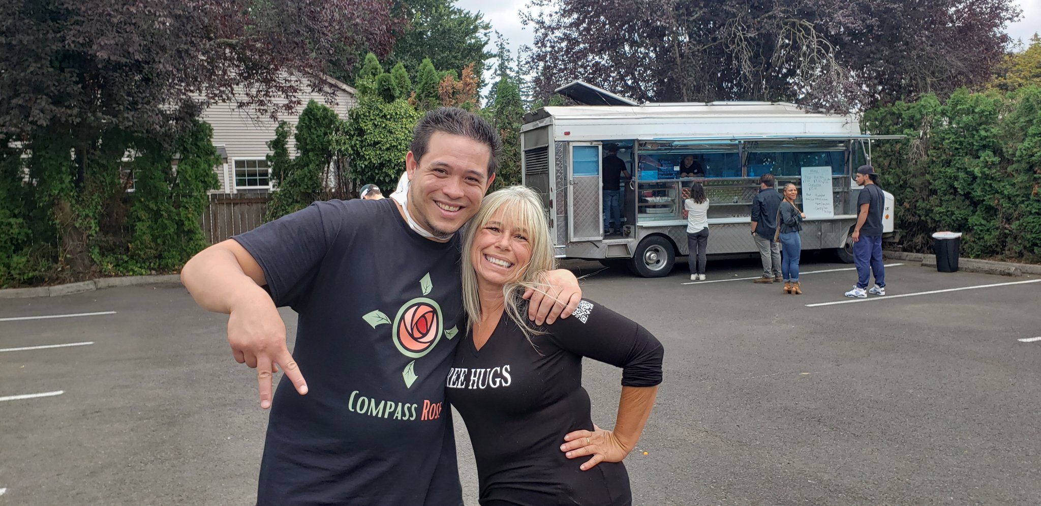Angel Prater in a FREE HUGS shirt with a community member in a Compass Rose shirt in a parking lot, a food truck behind them, with the Ascending Flow community