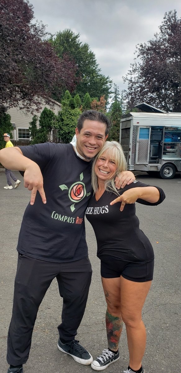 Angel Prater in a FREE HUGS shirt with a community member in a Compass Rose shirt, outdoors by a food trailer, with the Ascending Flow community