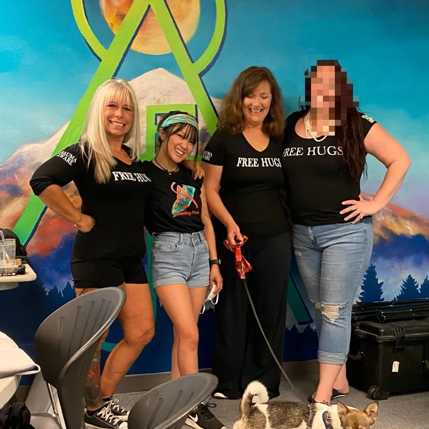 Angel Prater with three community members in FREE HUGS and Ascending Flow shirts in front of a mountain mural, a small dog on a leash in front