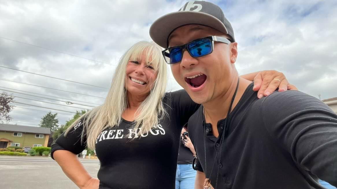 Selfie of Angel Prater in a FREE HUGS shirt with a member of the Ascending Flow crew in a cap and sunglasses, in a parking lot