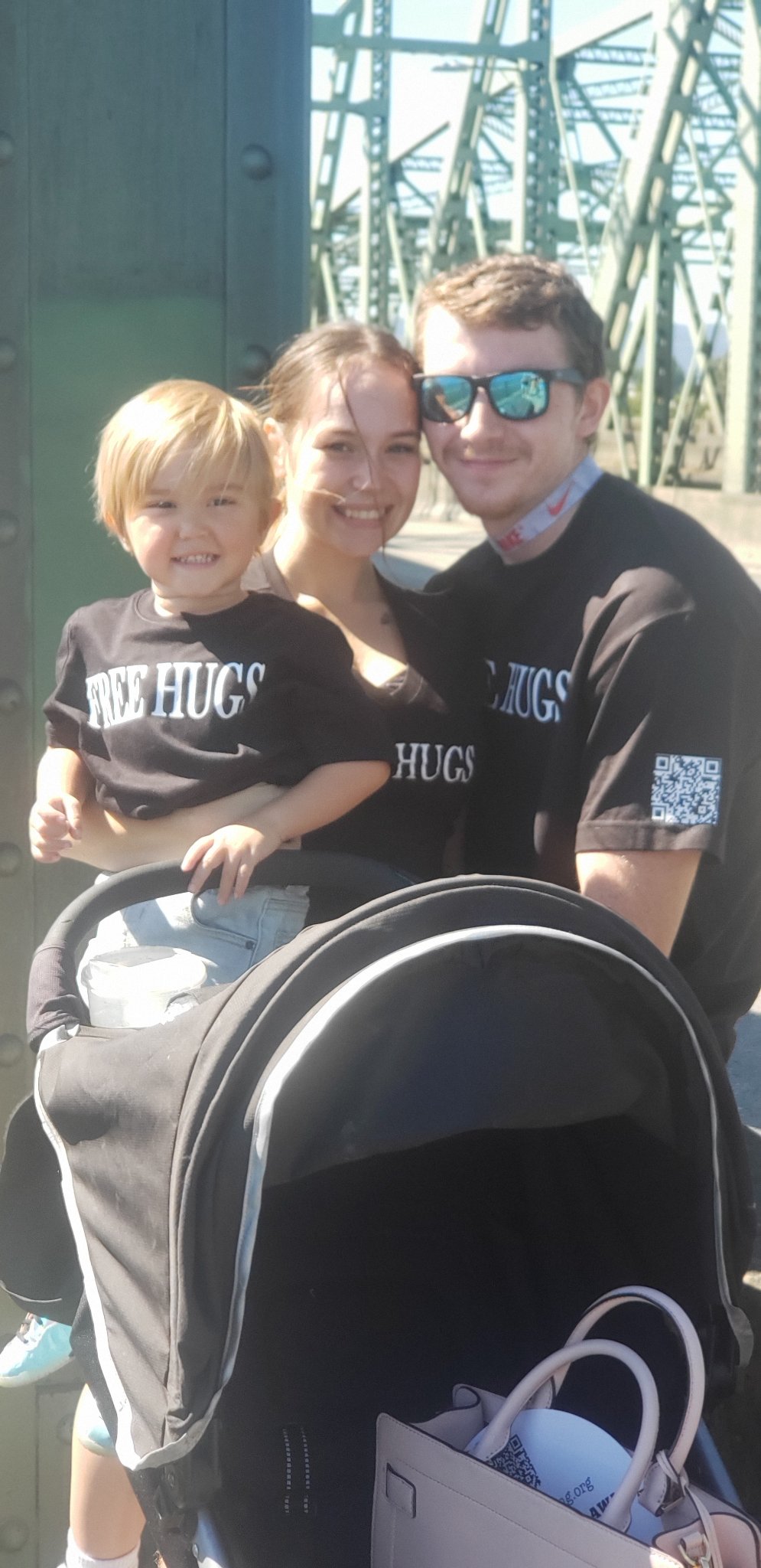 A crew member holding a young child in a FREE HUGS shirt on the bridge at Hands Across the Bridge, beside a fellow crew member in sunglasses, with a stroller in front