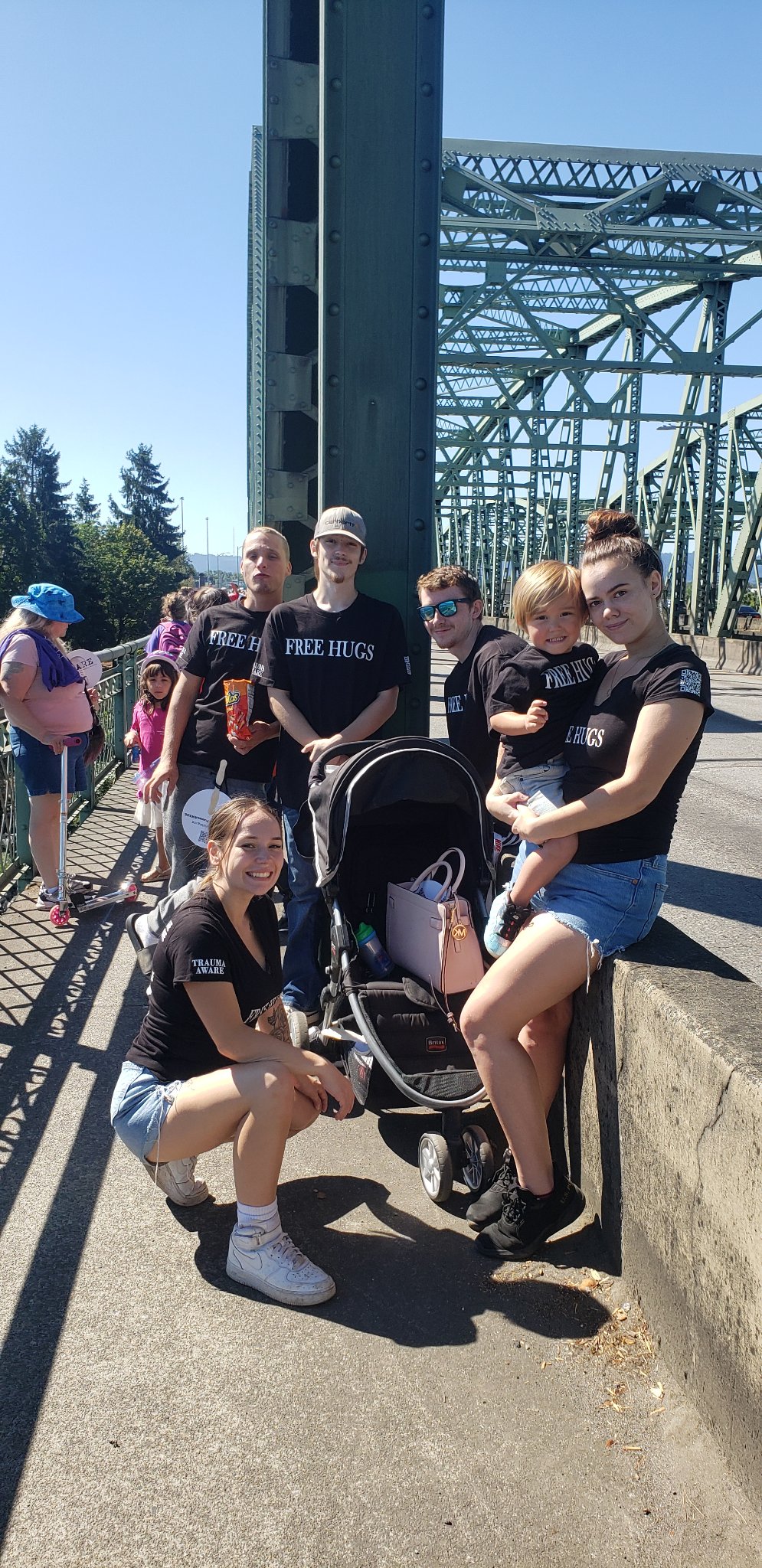 Free Hugs crew members in FREE HUGS shirts gathered on a green steel truss bridge at Hands Across the Bridge, with a stroller