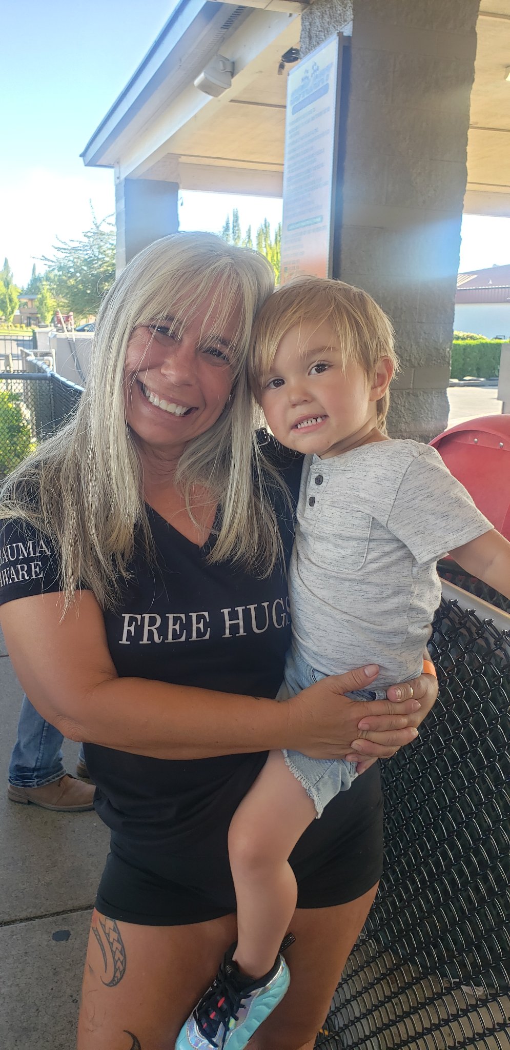 Angel Prater in a FREE HUGS shirt holding a young child under a pavilion at Hands Across the Bridge