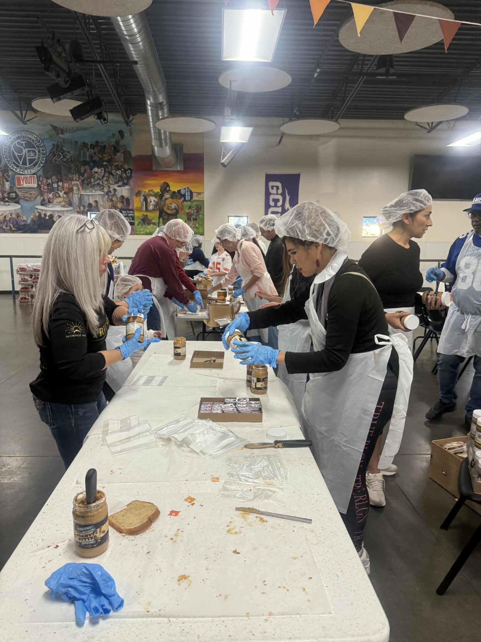 Volunteers in hairnets and aprons making peanut butter and jelly sandwiches along a serving table at St. Vincent de Paul, Angel Prater working the table in an Alchemize HOPE shirt