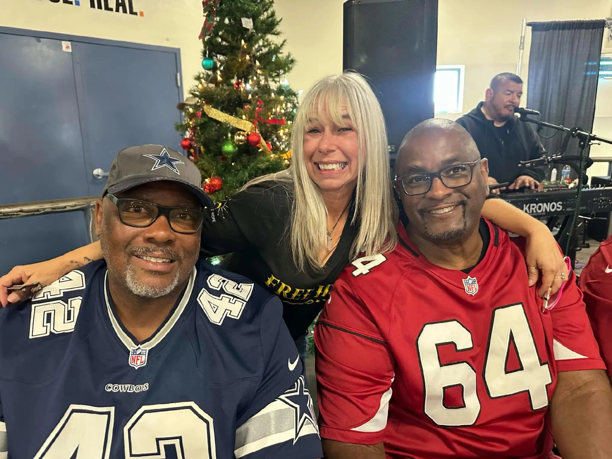 Angel Prater in a FREE HUGS shirt with two players by a Christmas tree at a Champions for the Homeless event