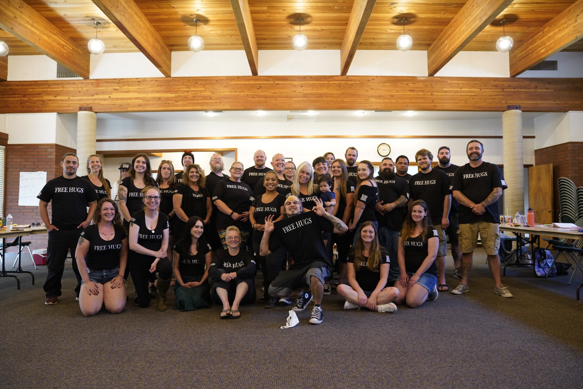 A large training class in a wood-beamed hall in Eugene, Oregon, nearly everyone wearing FREE HUGS shirts, Angel Prater at the center