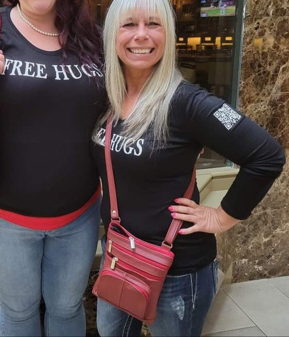 Angel Prater smiling in a FREE HUGS shirt beside a fellow hugger in a marble lobby