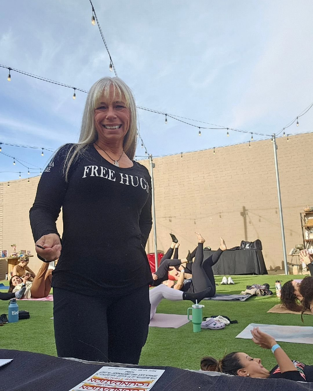 Angel Prater in a FREE HUGS shirt at an outdoor group yoga session on turf with string lights overhead