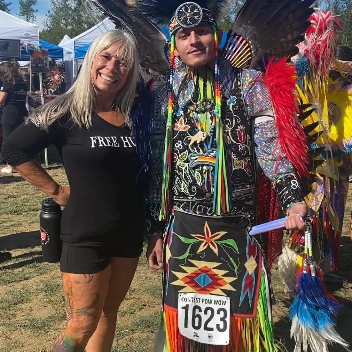 Angel Prater in a FREE HUGS shirt beside a powwow dancer in full regalia at a Native American and Indigenous event in Oregon
