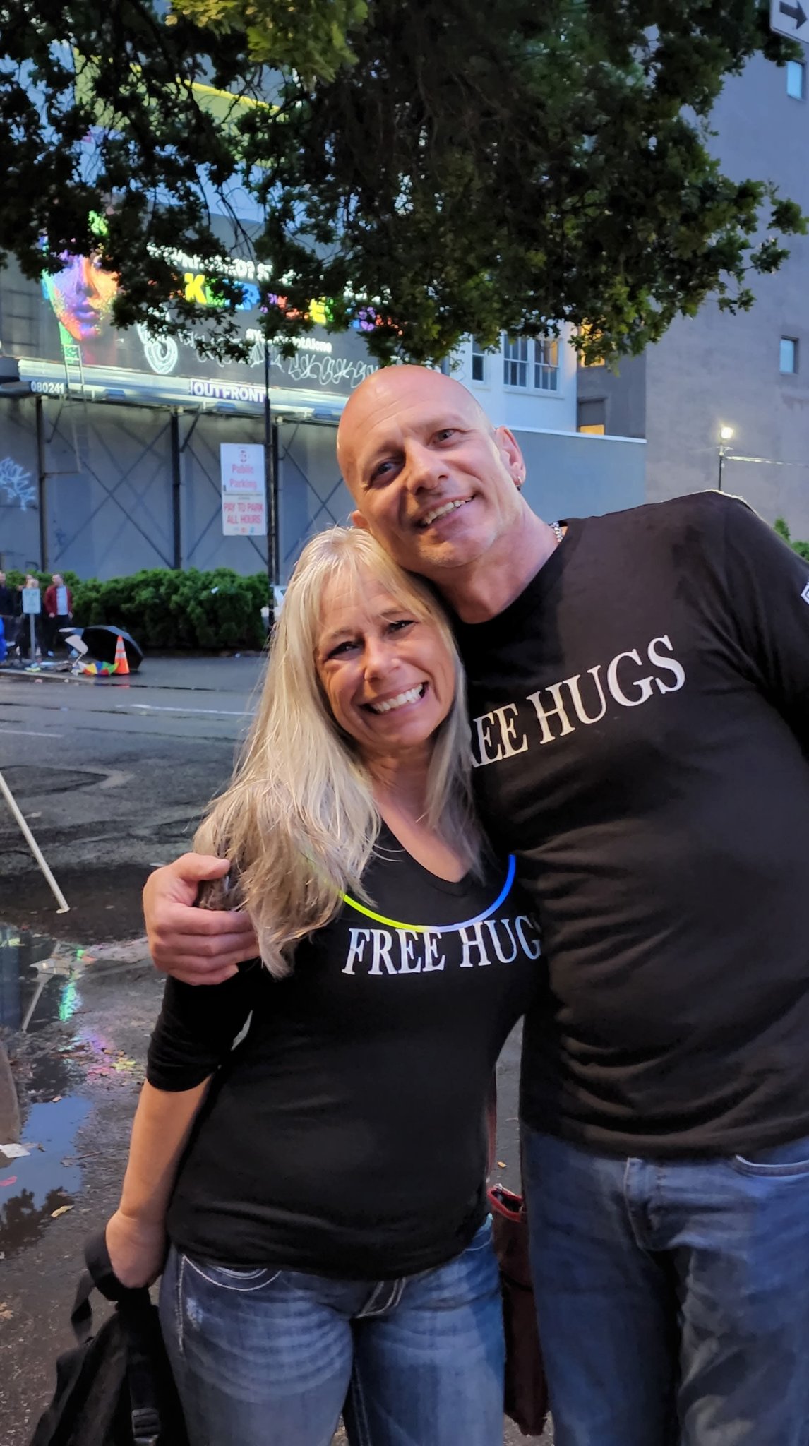 Angel Prater in a FREE HUGS shirt with a fellow hugger on a street at dusk at the CareOregon Starlight Parade
