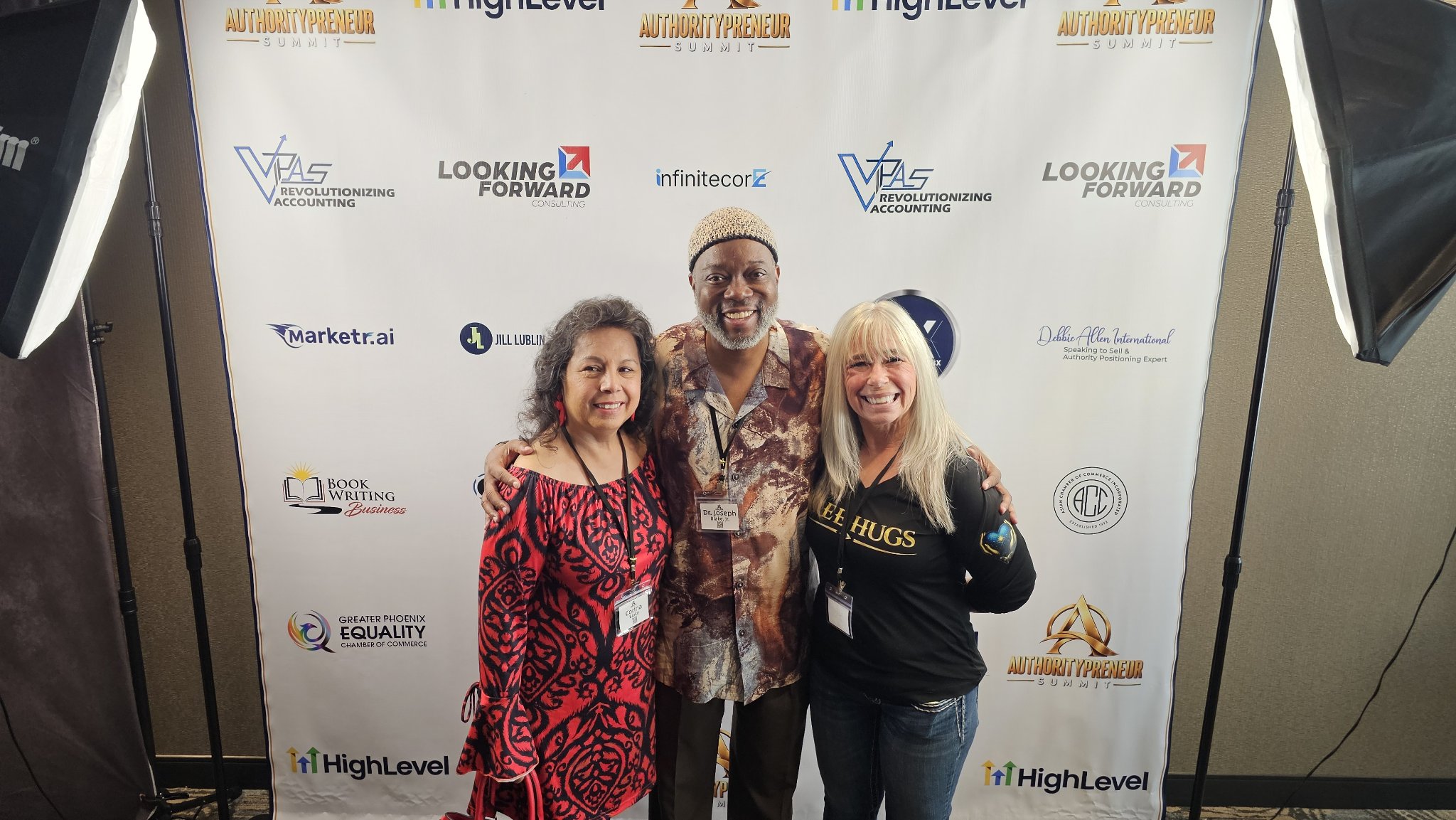 Angel Prater in a FREE HUGS shirt with two fellow attendees in front of an Authoritypreneur Summit backdrop