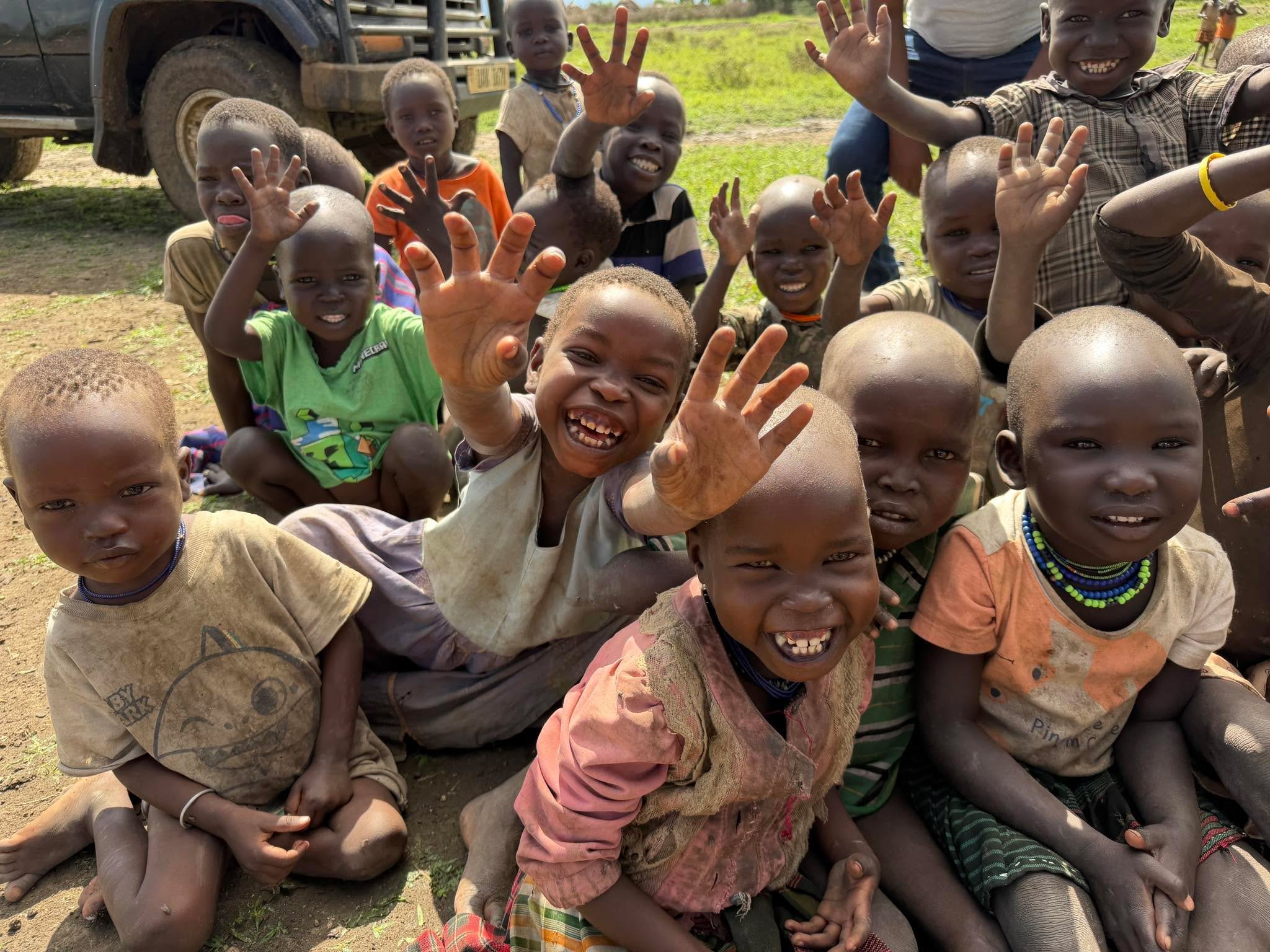 Children in Uganda waving joyfully at the camera, a vehicle behind them