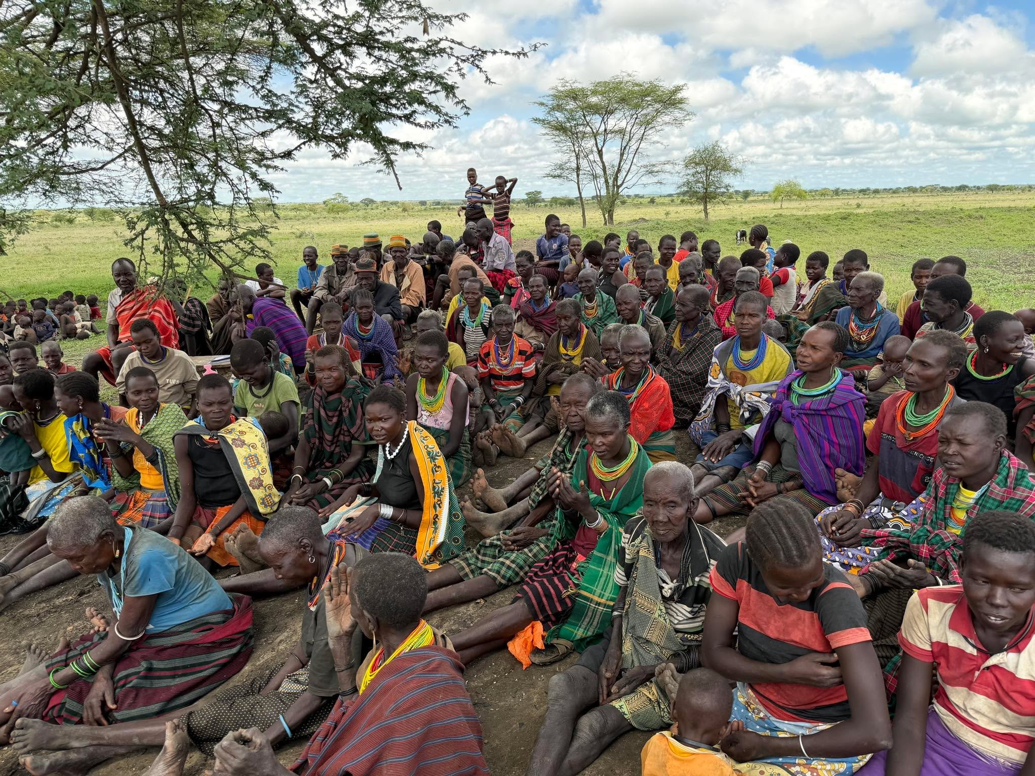 A wide view of a large community gathering seated outdoors under a tree in Uganda