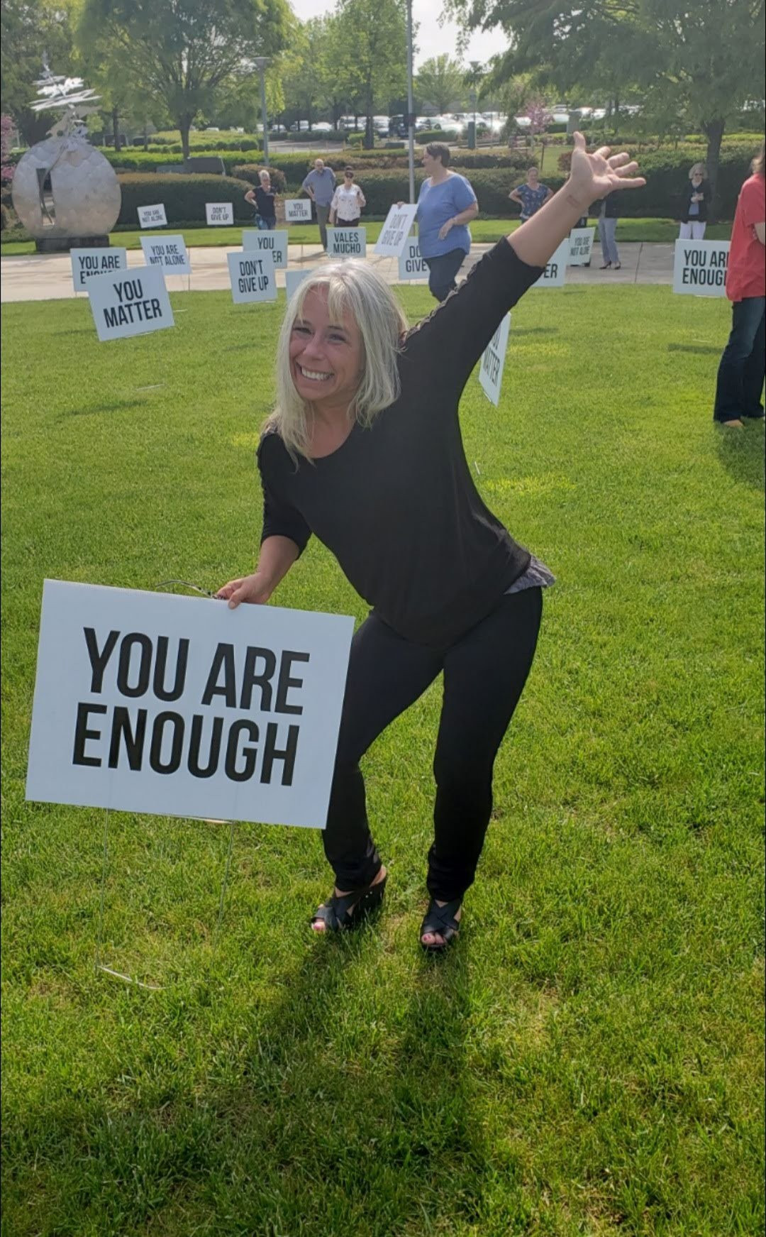 Angel Prater on a lawn holding a YOU ARE ENOUGH sign, with more signs behind her reading You Matter, Don't Give Up, Vales Mucho, and You Are Not Alone