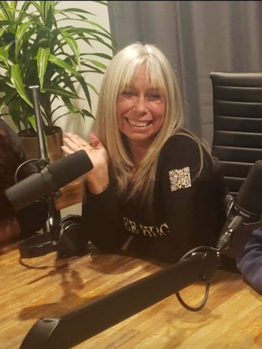 Angel Prater at a podcast studio microphone, wearing a FREE HUGS shirt