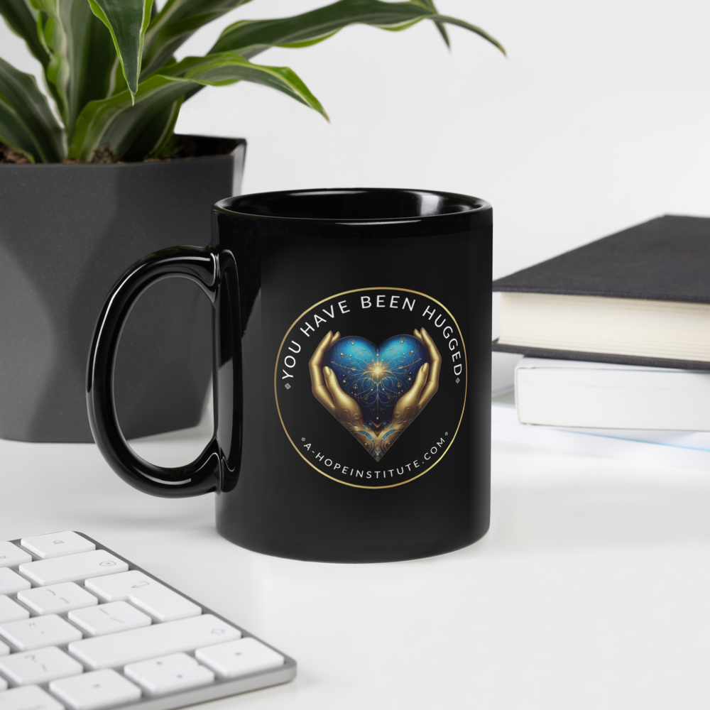 Black mug with the 'You Have Been Hugged' heart-and-hands emblem, on a desk beside a plant and books